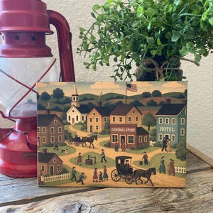 May include: A rectangular wooden sign depicting a folk art village scene with buildings, figures, and horse-drawn carriages. The sign includes the text "GENERAL STORE" and "HOTEL". A red lantern and a small green plant are in the background.