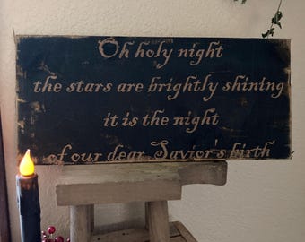 May include: A rustic wooden sign with the words "Oh holy night" in a decorative font. The sign is black with gold lettering and sits on a small wooden stool. A black candle with a faux flame and red berries are in the foreground.