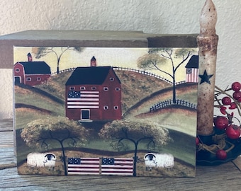 Peut inclure: Une pancarte en bois peinte représentant une grange rouge et une ferme avec des drapeaux américains sur la façade. La scène se déroule dans un paysage rural avec des collines et des arbres. Deux moutons blancs se trouvent au premier plan avec des drapeaux américains derrière eux.