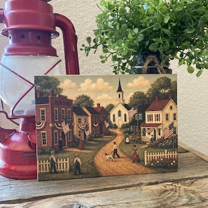 Peut inclure: Une impression rectangulaire représentant une scène de ville américaine pittoresque avec une église, des maisons et des personnages. L'œuvre présente une route de terre, des drapeaux américains et une lanterne rouge à gauche. L'ensemble est chaleureux, évoquant l'Amérique historique.