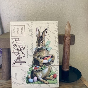 May include: Vintage postcard featuring a brown rabbit holding a basket of colorful Easter eggs. The postcard has a faded, antique look and includes the text "Postcard" and "I love you April".