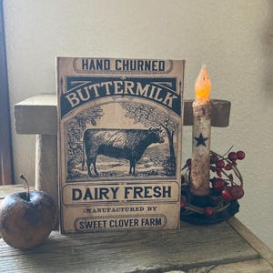 May include: A vintage-style label with the text "HAND CHURNED BUTTERMIK DAIRY FRESH MANUFACTURED BY SWEET CLOVER FARM" and a black and white illustration of a cow. The label is on a wooden surface with a small candle and red berries.