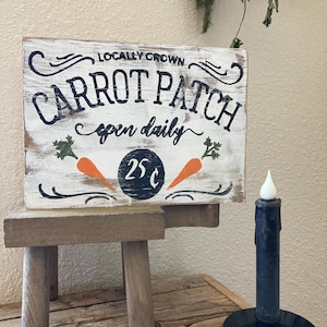 May include: A rustic wooden sign with the text "CARROT PATCH" and "open daily" in black lettering, featuring orange carrot illustrations. The sign is propped on a small wooden stool. A black candle sits to the right.