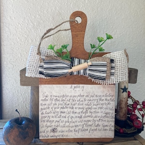 May include: A wooden cutting board with a black and white ribbon bow and a handwritten recipe for a "pottile py" on the front. The recipe includes instructions for making a potato and apple pie.