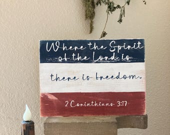 May include: A rectangular wooden sign with a distressed finish featuring horizontal stripes in navy blue, white, and red. The text reads: Where the Spirit of the Lord is there is freedom. 2 Corinthians 3:17 in white and dark blue script.