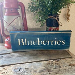 May include: A distressed blue wooden sign with the word "Blueberries" in white lettering. A red lantern and a small potted plant are in the background, all set on a rustic wooden surface.
