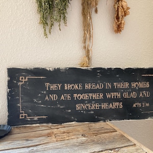 May include: A rustic, rectangular wooden sign with the text "THEY BROKE BREAD IN THEIR HOMES AND ATE TOGETHER WITH GLAD AND SINCERE HEARTS ACTS 2:46" in a distressed font. A candle sits in a black holder.