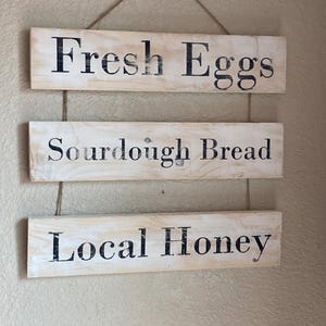 May include: A hanging wall sign consisting of three rectangular light-coloured wooden planks connected by twine. Each plank features black text: the top reads Fresh Eggs, the middle reads Sourdough Bread, and the bottom reads Local Honey.