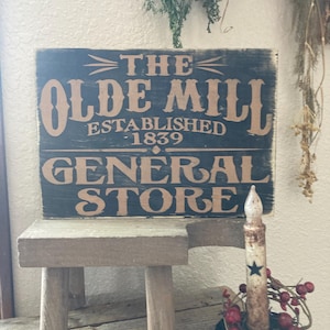 May include: A black wooden sign with white and brown lettering reads "The Olde Mill Established 1839 General Store". The sign is propped up on a small wooden stool. A white candle with a black star sits in a black metal candle holder with red berries.