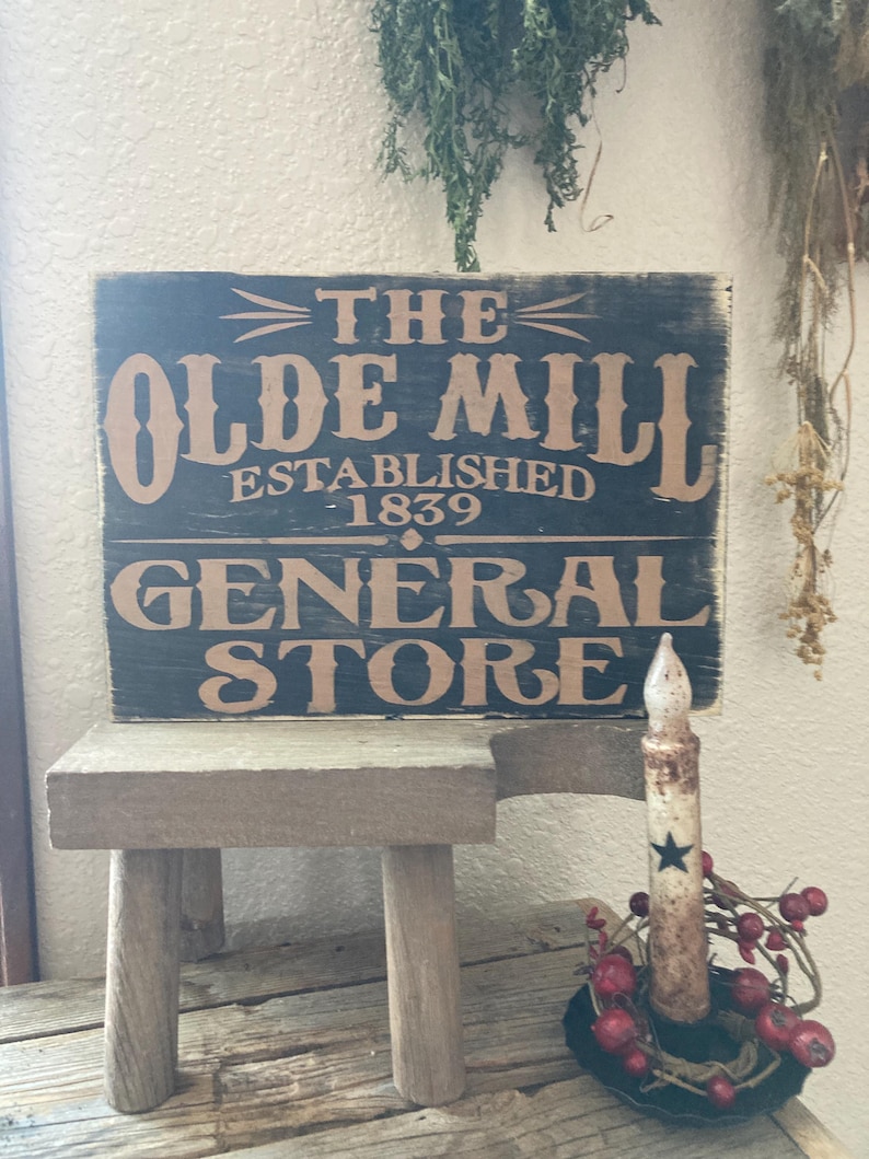 May include: A black wooden sign with white and brown lettering reads "The Olde Mill Established 1839 General Store". The sign is propped up on a small wooden stool. A white candle with a black star sits in a black metal candle holder with red berries.