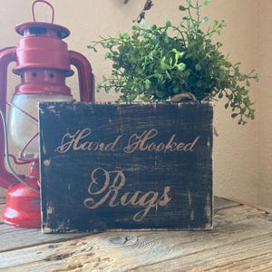 May include: A rectangular wooden sign with a distressed black finish and the text Hand Hooked Rugs in a light-colored script font. The sign is positioned next to a bright red metal lantern and a small green potted plant with delicate leaves.
