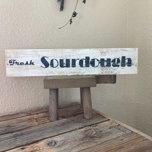 Peut inclure: Panneau en bois rectangulaire avec l'inscription "Fresh Sourdough" en lettres noires sur fond blanc vieilli. Le panneau est posé sur un petit tabouret en bois, devant une table en bois rustique.