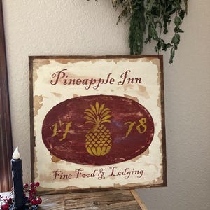 May include: Wooden sign with the text "Pineapple Inn" and "Fine Food & Lodging". The sign features a maroon oval with a gold pineapple and the numbers "17" and "78". The sign has a distressed, vintage look.