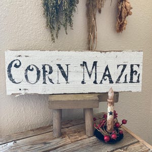 May include: A rustic wooden sign with the words "Corn Maze" in black lettering. The sign is propped up on a small wooden stand, with a candle and red berries in front. Dried herbs hang in the background.