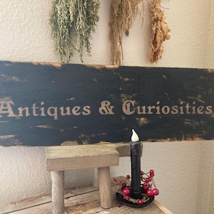Peut inclure: Panneau en bois noir vieilli avec l'inscription "Antiquités & Curiosités". Une bougie noire avec une couronne de baies rouges est posée sur un petit tabouret en bois. Des herbes séchées sont suspendues au-dessus.