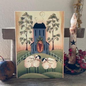 May include: A hand-painted wooden folk art sign featuring a blue house with a red heart on the front, two white sheep with green foliage, and two trees in the background.