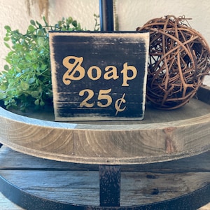 May include: A black wooden sign with white distressed paint and gold lettering that reads "Soap 25¢". The sign is sitting on a wooden tiered tray.