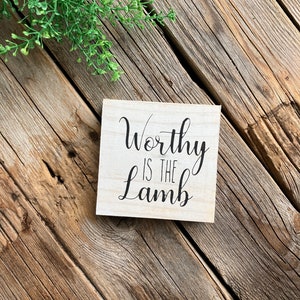 May include: A wooden sign with the text "Worthy is the Lamb" in black cursive lettering.