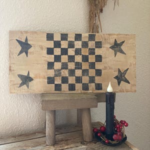 May include: A rustic wooden sign with a checkerboard pattern and four star designs. The sign is displayed on a small wooden stool. A black candle with a red berry wreath sits nearby, creating a charming, country-style aesthetic.