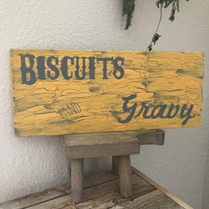 May include: A distressed wooden sign, painted yellow with the words "BISCUITS and Gravy" in dark grey. The sign rests on a small, rustic wooden stool. The overall aesthetic suggests a country kitchen or farmhouse style.