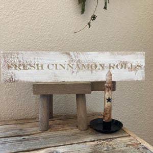 May include: A rustic wooden sign with the words "FRESH CINNAMON ROLLS" in brown lettering. The sign rests on a small wooden stand. A candle with a black star is in a black dish.