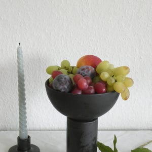 Può includere: Scodella in ceramica nera con un piedistallo, piena di uva, prugne e una pesca. Una candela blu chiaro attorcigliata si trova in un candelabro nero su una tovaglia bianca.