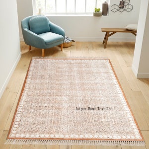 May include: A light brown area rug with a white geometric pattern and a white fringe border. The rug is on a wooden floor and the text "Jaipur Home Textiles" is printed in the center of the rug.