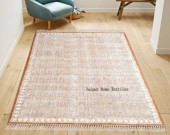 May include: A light brown area rug with a white geometric pattern and a white fringe border. The rug is on a wooden floor and the text "Jaipur Home Textiles" is printed in the center of the rug.