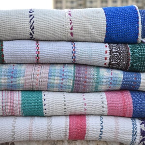 Pode incluir: Uma pilha de cinco colchas enroladas. Cada colcha apresenta uma base branca com detalhes coloridos, incluindo azul, rosa, verde e roxo. As colchas têm vários padrões costurados e bordas decorativas, mostrando um design de patchwork.