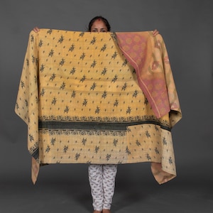 May include: A large, rectangular textile with a yellow background and a repeating black floral pattern. The textile has a dark green border and a pink floral patterned section on the right side. The textile is being held up by a person.
