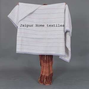 May include: A white striped throw blanket with blue and red stripes. The blanket is held up by a person wearing a brown and gold patterned sari. The text "Jaipur Home textiles" is printed on the blanket.