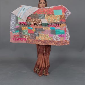 Puede incluir: Una colcha de retazos colorida con una variedad de telas y patrones. La colcha está sostenida por una persona que lleva un sari con estampado marrón y blanco. El texto "Jaipur Home textiles" es visible en la colcha.