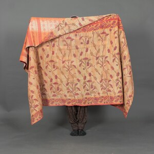 Peut inclure: Un châle beige et orange à motifs floraux. Le châle a une bordure de motifs floraux rouges et oranges. Le châle est drapé sur une personne, dont seules les jambes et les pieds sont visibles, portant un pantalon marron à motifs et des chaussures noires.