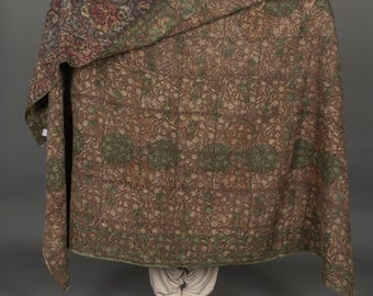 May include: A large, patterned quilt with a brown and green floral design. The quilt is draped, revealing a portion of the pattern and the person holding it. The quilt's colors include shades of brown, green, and red.