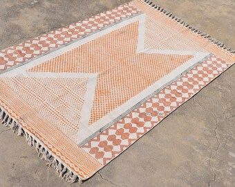 May include: A white and orange handwoven rug with a geometric pattern. The rug has a fringe border and is made of cotton.