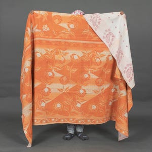 May include: An orange floral patterned blanket with a white and pink paisley patterned scarf. The blanket has a repeating pattern of roses and leaves. The scarf has a white background with pink paisley and dot designs. The blanket and scarf are being held up.