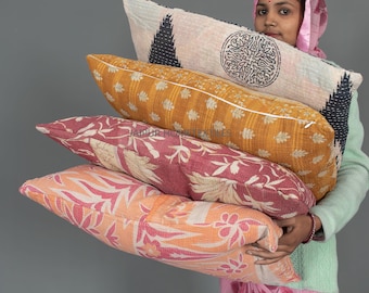 May include: A woman in a pink headscarf holds a stack of four colorful patterned throw pillows. The pillows are made of fabric with intricate designs and are stacked in a pyramid shape.