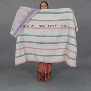 May include: A white kantha quilt with colorful stripes and a purple border. The quilt is held up by a person wearing a brown and gold sari. The text "Jaipur Hometextiles" is visible on the quilt.