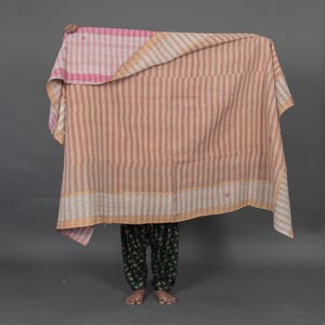 May include: A large, reversible blanket with a pink and white plaid pattern on one side and vertical stripes in shades of tan and pink on the other. The blanket is held up, partially obscuring the person behind it. The blanket's edges are trimmed with a gold border.