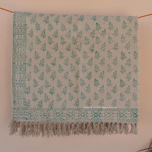 Op de afbeelding: Een wit katoenen vloerkleed met een groen bloemenpatroon en franjes aan de randen. Het vloerkleed hangt aan een muur met een touw dat aan elke hoek is bevestigd. De tekst "Image © Jaipur Hometextiles" is gedrukt in de rechterbenedenhoek van het vloerkleed.