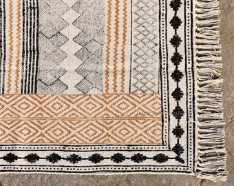May include: Close-up of a woven rug with a geometric pattern in shades of brown, black, and white. The rug has a fringe border.