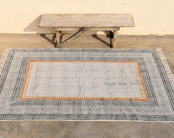 Puede incluir: Una alfombra blanca y negra con estampado, borde marrón y flecos. La alfombra está colocada sobre un suelo de hormigón con un banco de madera en el fondo. El texto "Jaipur Home Textiles" está impreso en la alfombra.