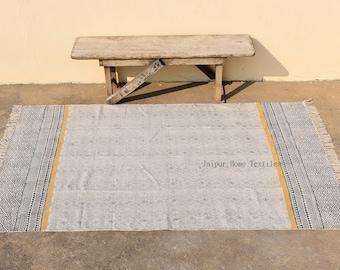 Puede incluir: Una alfombra rectangular gris claro con un borde geométrico en blanco y negro y una franja amarilla. La alfombra tiene un borde con flecos y está colocada sobre una superficie de hormigón. Un banco de madera está en el fondo.