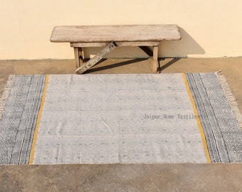 Puede incluir: Una alfombra gris y blanca con estampado, borde amarillo y flecos. La alfombra está colocada sobre un suelo de hormigón con un banco de madera en el fondo. El texto "Jaipur Home Textiles" está impreso en la alfombra.