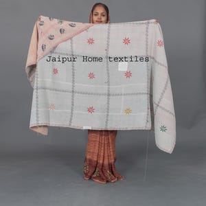 Peut inclure: Une courtepointe kantha blanche avec un motif floral rose sur un côté. La courtepointe est pliée en deux et tenue par une personne. La courtepointe a un motif en grille avec des fleurs brodées et une bordure de petits cercles. Le texte "Jaipur Home textiles" est visible sur la courtepointe.