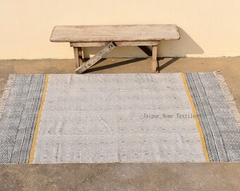 Puede incluir: Una alfombra rectangular gris claro con un borde geométrico en blanco y negro y una franja amarilla. La alfombra tiene un borde con flecos y está colocada sobre una superficie de hormigón. Un banco de madera está en el fondo.