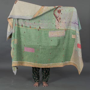 Könnte beinhalten: Ein Patchwork-Quilt in Grün-, Beige- und Pastelltönen. Der Quilt weist eine Vielzahl von genähten Mustern und Flicken auf. Der Quilt wird hochgehalten und zeigt die Unterschenkel und Füße einer Person, die eine dunkelgrüne Hose mit Muster trägt.