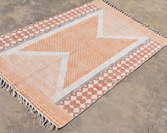 May include: A white and orange handwoven rug with a geometric pattern. The rug has a fringe border and is made of cotton.