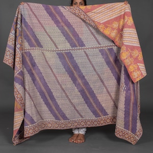 Op de afbeelding: Een grote, omkeerbare deken met een streepjespatroon in tinten paars, beige en oranje. De deken heeft aan één kant een bloemenpatroon en een geometrische rand. De deken wordt door een persoon vastgehouden.
