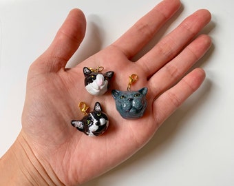 Puede incluir: Tres pequeños dijes con forma de cabeza de gato con cierres de langosta dorados. Un dije es gris sólido, uno es blanco y negro con hocico blanco, y otro es blanco y negro con una mancha blanca en la nariz y la frente, todos con detalles pintados.